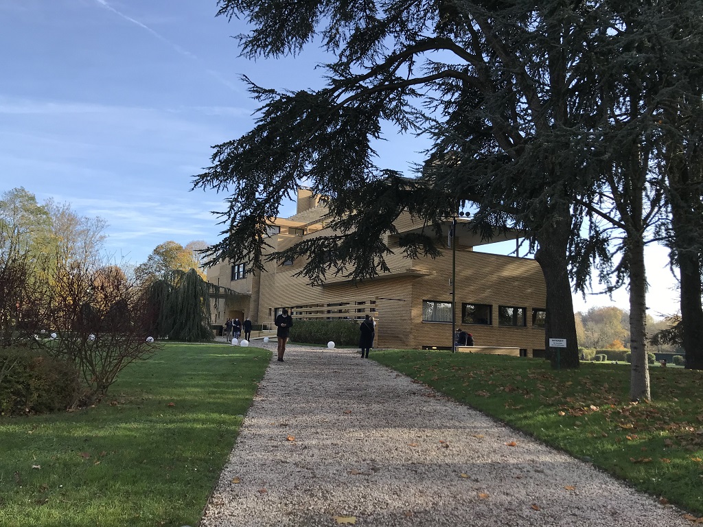 Ein Kiesweg führt zu einem Haus aus Ziegelsteinen. Rechts und links des Weges befindet sich Rasen. Vor dem Haus steht eine große Zeder. Auf dem Kiesweg sind Menschen zu sehen