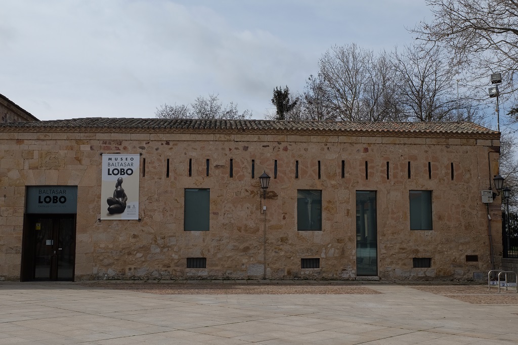 Das Foto zeigt das Museum Baltasar Lobo, ein langgestrecktes Gebäude aus groben, unregelmäßigen Steinen und einem Dach aus grauen Dachziegeln aus Ton. Auf der linken Seite ist der Eingang. Auf der Fassade hängt ein Plakat, auf dem eine Figur des Künstlers abgebildet ist. Die Figur hat kräftige Oberschenkel.