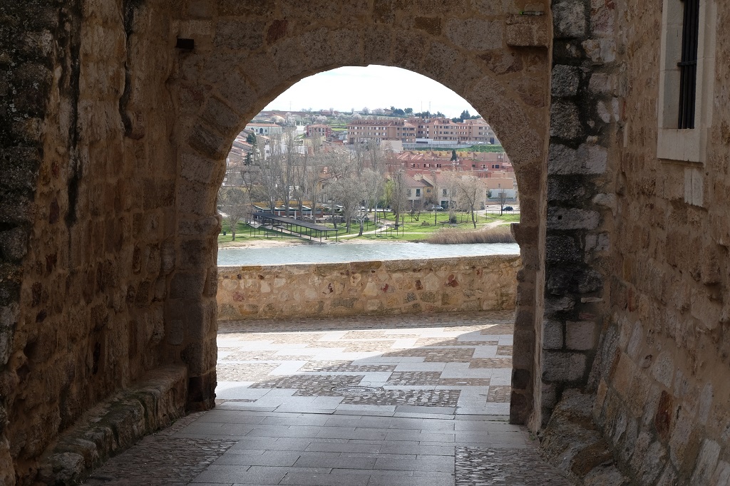 Hinter einem dunklen Torbogen erscheint ein schmaler Streifen des Duero sowie das gegenüberliegende Ufer. Der Boden hinter dem Tor ist abwechselnd mit Platten und Kopfsteinpflaster bedeckt.