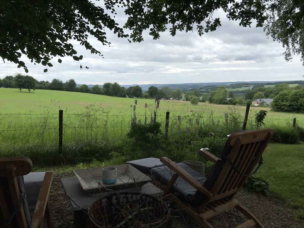 Das Foto zeigt zwei Holzliegestühle mit einem Tisch dazwischen. Sie sind auf eine Wiese auf dem Nachbargrundstück ausgerichtet. Im Hintergrund ist eine leicht gewellte Landschaft aus Wiesen und Wäldern zu sehen.